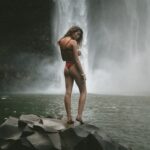 woman in red panty and bra standing of black stone near waterfalls